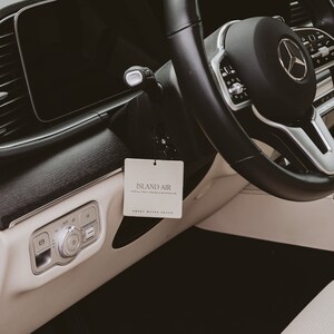 May include: Interior shot of a car's dashboard and steering wheel. A white rectangular air freshener with the text "ISLAND AIR" hangs from the rearview mirror. The steering wheel is black with a silver Mercedes-Benz logo.