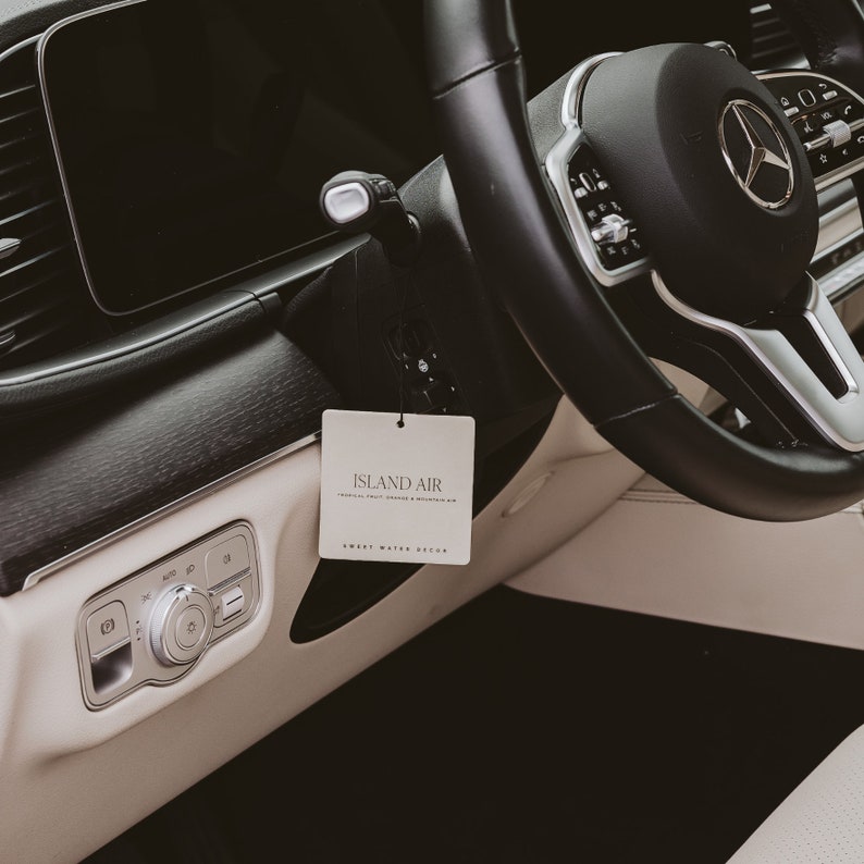 May include: Interior shot of a car's dashboard and steering wheel. A white rectangular air freshener with the text "ISLAND AIR" hangs from the rearview mirror. The steering wheel is black with a silver Mercedes-Benz logo.