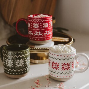 May include: Three ceramic mugs featuring festive patterns. One red mug with white reindeer and snowflakes, one dark green mug with white snowflakes, and one white mug with red and green snowflakes. The white mug is filled with whipped cream.