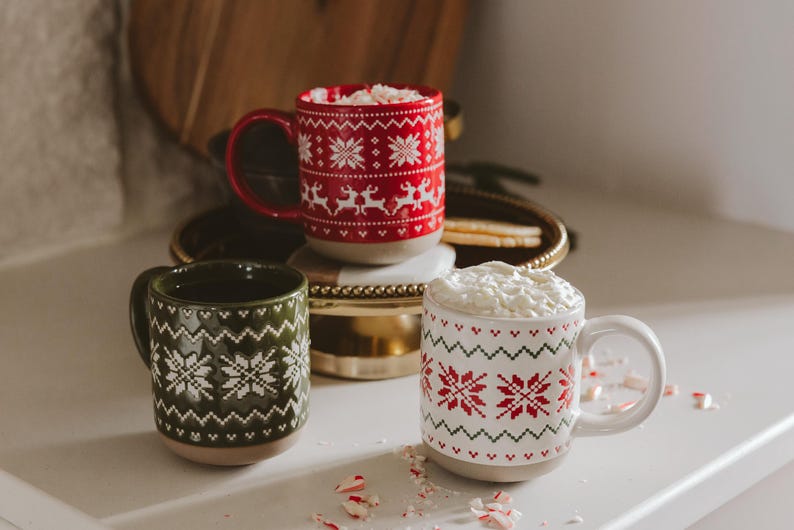 May include: Three ceramic mugs featuring festive patterns. One red mug with white reindeer and snowflakes, one dark green mug with white snowflakes, and one white mug with red and green snowflakes. The white mug is filled with whipped cream.