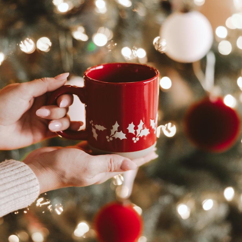 May include: A red ceramic mug with a white holly design is held in front of a Christmas tree. The mug has a handle and a light-coloured base. The background features blurred Christmas lights and baubles.