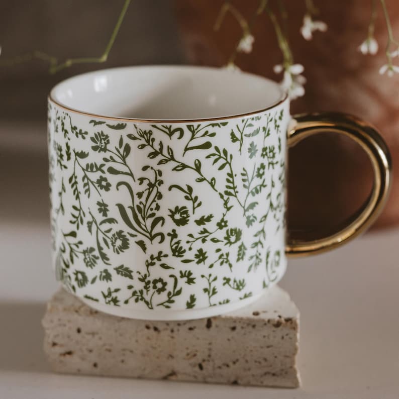 May include: White ceramic mug with a gold handle and rim, adorned with a green floral pattern. The mug is resting on a small, light-colored stone. The design features delicate leaves and flowers.