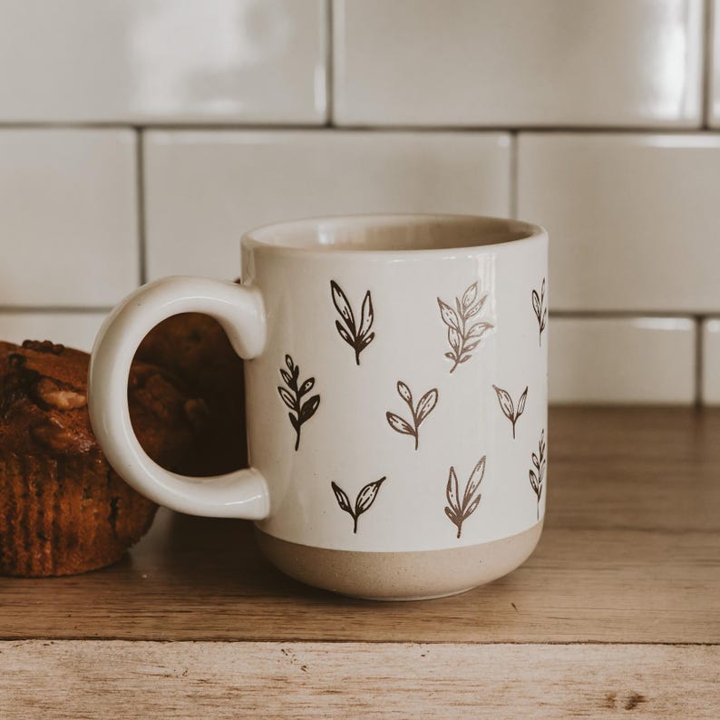 May include: A white ceramic mug with a beige base and a large handle. The mug is decorated with a pattern of black leaf illustrations. A muffin is visible to the left of the mug, on a wooden surface.