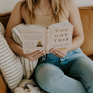 May include: A woman is reading a book titled "You Got This: 52 Devotions of Encouragement for Empowering Hardworking Women" by Crystal Dorsey. The book cover is pink with gold lettering. The woman is wearing a light brown tank top and blue jeans.
