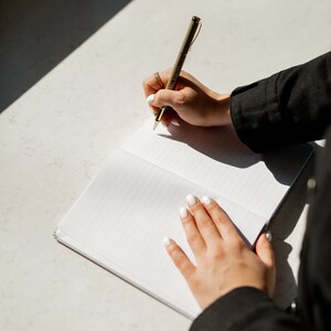 May include: A person writing in a blank notebook with a gold pen. The notebook is open and has lined pages. The person is wearing a black shirt and has white nail polish.