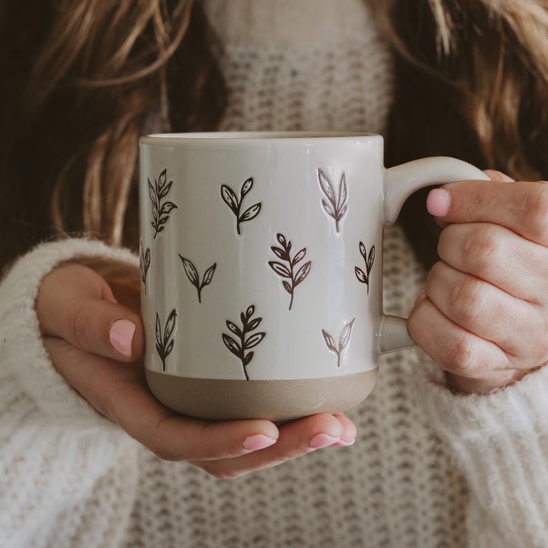 May include: Cream-colored ceramic mug with a brown base and handle, adorned with dark brown leaf illustrations. The mug is held by a person wearing a white sweater. The mug is designed for hot beverages.