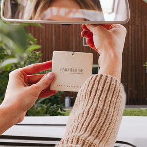 May include: A rectangular, light brown air freshener with the word "FARMHOUSE" printed on it, held up near a car's rearview mirror. The text "APPLE, CLOVE, CINNAMON & ORANGE" and "SWEET WATER DEC" are also visible. The freshener is suspended by a black string.