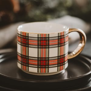 May include: A ceramic mug with a white base and a red, black, and green plaid pattern. The mug has a gold-colored handle and rim. The mug is sitting on a black plate.