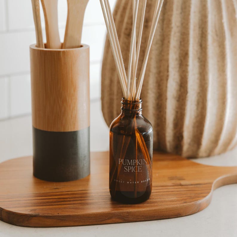 May include: A brown glass reed diffuser bottle with the text PUMPKIN SPICE printed on the front, containing several thin wooden reeds. Next to it is a cylindrical wooden utensil holder with a dark base and three light wooden kitchen spoons.