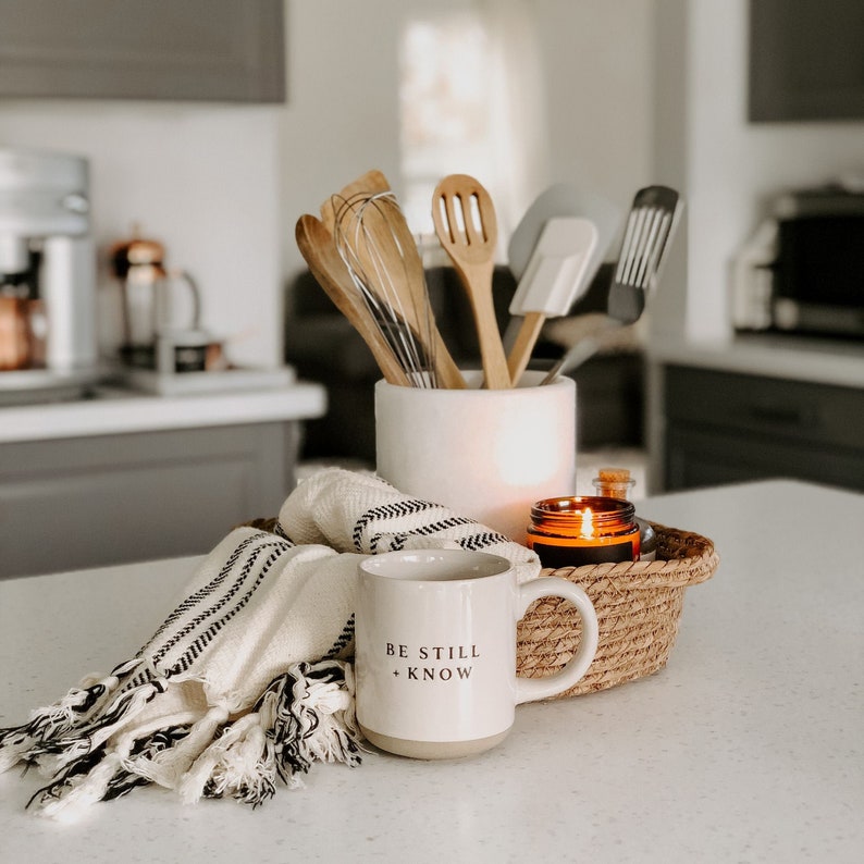 May include: A kitchen countertop display featuring a white mug with "BE STILL + KNOW" text, a woven basket with a lit candle, a folded black and white striped towel, and wooden kitchen utensils in a white ceramic holder.