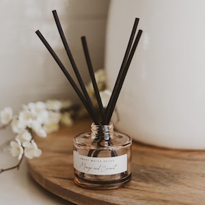 May include: A clear glass diffuser bottle with black reeds. The bottle has a label that reads "Sweet Water Decor Mango and Coconut." The diffuser sits on a wooden tray, with white flowers in the background. Home decor.