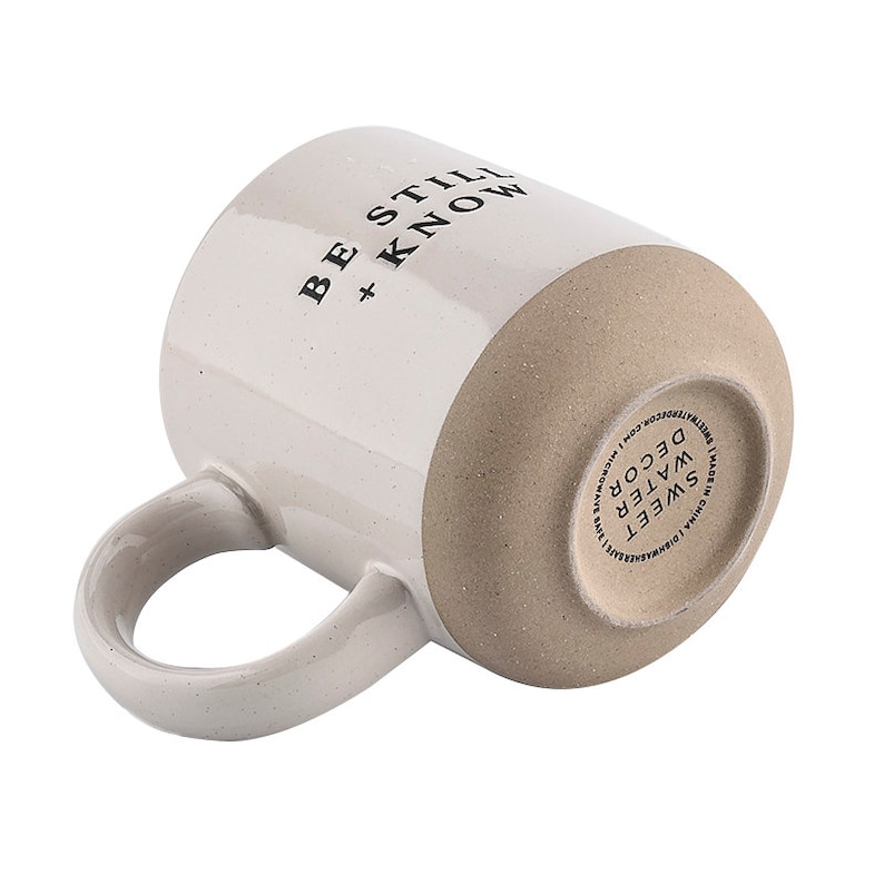 May include: Cream-colored ceramic mug with the text "BE STILL + KNOW" in black. The bottom of the mug is a light brown color. The mug has a curved handle and is designed for hot beverages. The mug is a part of the Sweet Water Decor collection.
