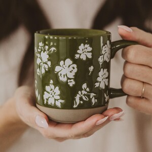May include: A green ceramic mug with white floral designs. The mug has a handle and a tan base. The floral pattern is embossed on the mug's surface. The mug is held by a person.