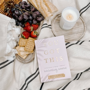 May include: A pink book with the title "You Got This" and the subtitle "90 Devotions to Equip + Empower hardworking women" by Melissa Horvath. The book is on a white and black striped blanket with a candle, grapes, crackers, and bread in the background.