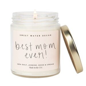 May include: A glass jar candle with a gold lid. The candle is white and has a pink label that says "SWEET WATER DECOR" and "best mom ever!" in black script. The label also says "SEA SALT, JASMINE, WOOD & CREAM" and "Made in the USA".