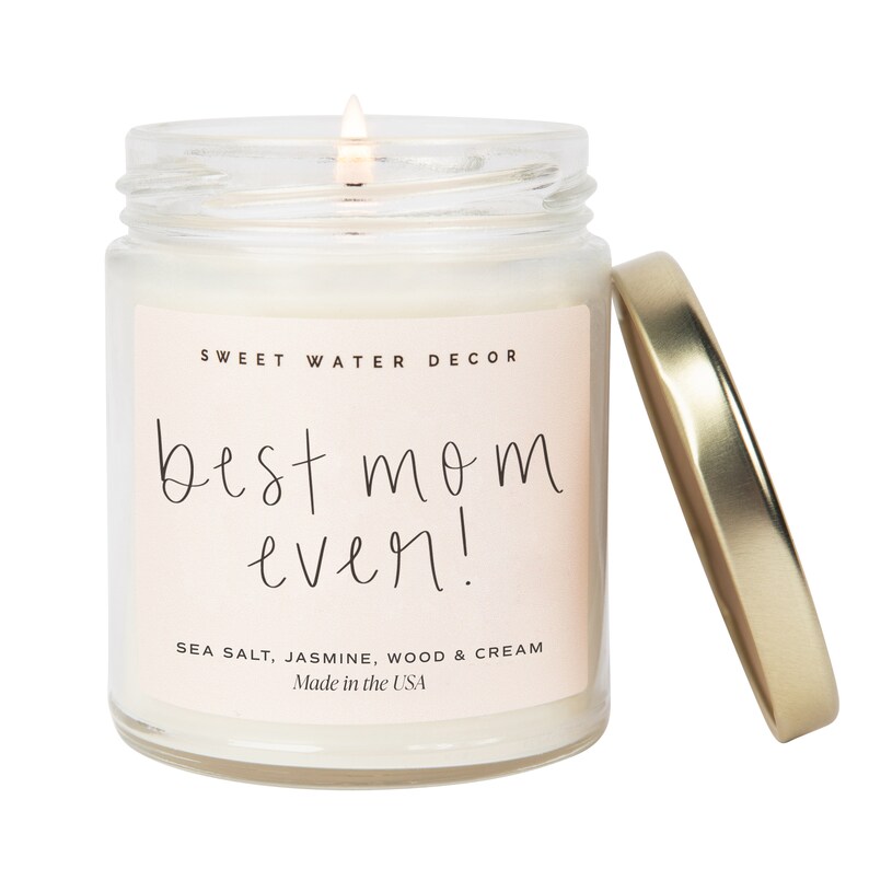 May include: A glass jar candle with a gold lid. The candle is white and has a pink label that says "SWEET WATER DECOR" and "best mom ever!" in black script. The label also says "SEA SALT, JASMINE, WOOD & CREAM" and "Made in the USA".