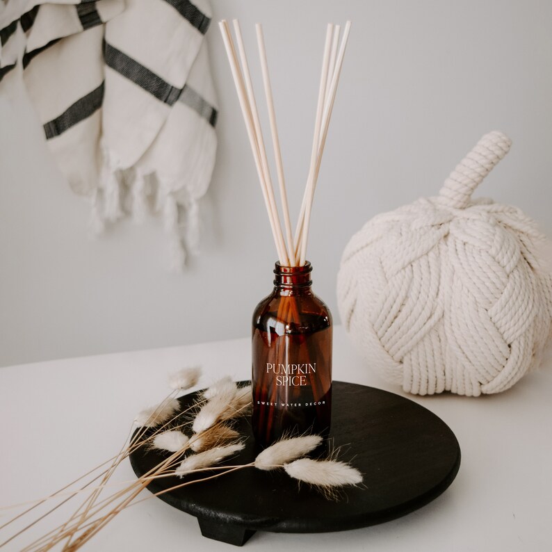 May include: An amber glass reed diffuser bottle with a label reading PUMPKIN SPICE SWEET WATER DECOR. The bottle contains several light-colored wooden reeds and is placed on a round black wooden tray alongside dried beige bunny tail grass stems.