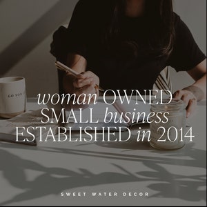 May include: A person writing with a pen on a desk with the text "woman OWNED SMALL business ESTABLISHED in 2014". A mug with "GO FOR IT" is on the left. A lit candle sits on the right. The brand name "SWEET WATER DECOR" is at the bottom.