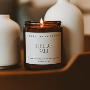 May include: A brown glass jar candle with a white label featuring the text SWEET WATER DECOR, HELLO FALL, CIDER, NUTMEG, CINNAMON & CLOVES, and Poured in the USA. The candle has a lit wick and is positioned between two white ceramic vases.