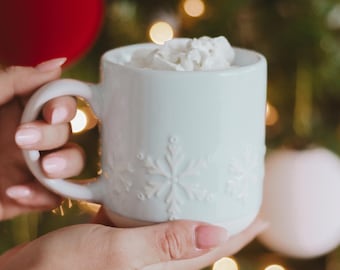 May include: A light blue ceramic mug with snowflake designs, filled with whipped cream. The mug is held by a person. The background features a Christmas tree with ornaments and lights, creating a festive holiday scene.