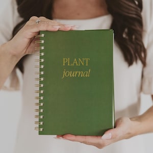 May include: A dark green spiral-bound journal with gold-colored wire binding. The front cover features the text "PLANT journal" printed in gold lettering. The journal has a solid matte finish and a rectangular shape with rounded corners.