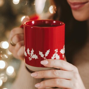 May include: A red ceramic mug with a white holly leaf pattern. The mug is held by a person. The mug has a beige base and a red handle. The background is blurred with bokeh lights.