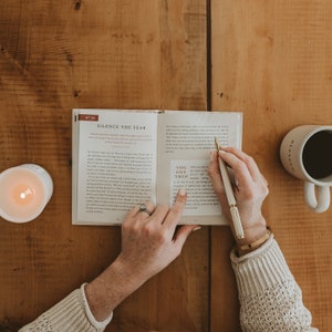 May include: A person reading a book with the text "You Got This!" highlighted. The book is open to a page with the title "Silence the Fear". A white pen is in the person's hand. A white mug with black coffee is on a wooden table.