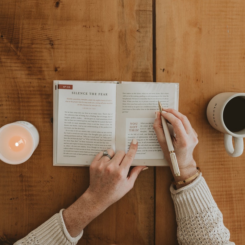 May include: A person reading a book with the text "You Got This!" highlighted. The book is open to a page with the title "Silence the Fear". A white pen is in the person's hand. A white mug with black coffee is on a wooden table.