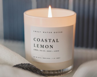 May include: A white candle with a frosted glass jar and a white label that reads "SWEET WATER DECOR COASTAL LEMON LEMON + SEA AIR + WOODS + JASMINE NET WT 237 g | MADE IN USA | 11 OZ CANDLE". The candle is lit and the flame is visible.