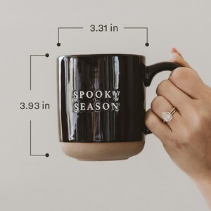 May include: A black ceramic mug with the words "SPOOKY SEASON" in white lettering. The mug is 3.93 inches tall and 3.31 inches wide. The bottom of the mug is a light brown color. The mug is held by a person.