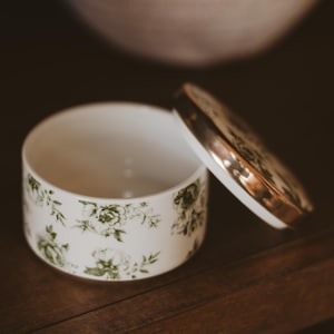 May include: A white ceramic trinket box with a lid, featuring a floral pattern in green. The lid has a gold-colored rim. The box is open, revealing its interior. The background is a dark wooden surface.