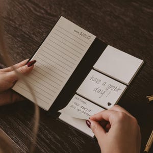 May include: A black notepad with lined paper and sticky notes. The notepad is open, revealing a page with the date 5/10/25 and lined paper. Sticky notes say "have a great day!" and "Don't forget... call dentist, pick up grocer."