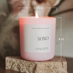 May include: A pink candle with a white label that reads "SWEET WATER DECOR XOXO ORANGE/TROPICAL FRUIT/MOUNTAIN AIR". The candle is 4 inches tall and 3.25 inches wide.