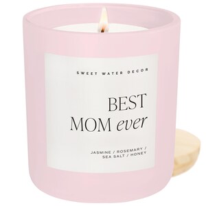 May include: A pink glass candle jar with a white label that reads "BEST MOM ever" and "SWEET WATER DECOR". The candle has a white wax and a lit wick. The label also lists the scent as "Jasmine / Rosemary / Sea Salt / Honey".