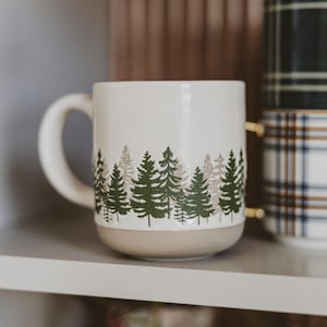 May include: A cream-colored ceramic mug featuring a wrap-around forest pattern of dark green and light beige pine trees. The mug has a smooth, rounded handle and a solid, unglazed beige base, providing a clean and simple design for daily use.