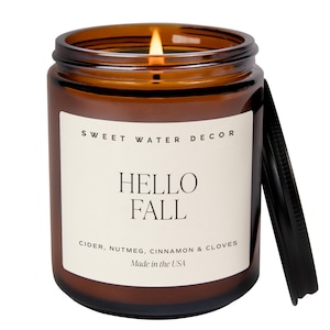May include: A lit candle in an amber glass jar with a black lid. The white label reads SWEET WATER DECOR, HELLO FALL, CIDER, NUTMEG, CINNAMON & CLOVES, and Made in the USA. The candle features a single burning wick centered in the wax.