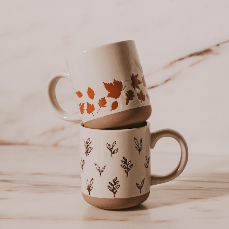 May include: Two stacked ceramic mugs. The top mug has orange leaf designs, while the bottom mug features brown leaf and sprig illustrations. Both mugs have a cream-colored body with a tan base and handle.