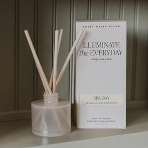 May include: A frosted glass reed diffuser bottle with light-colored reeds and a matching white rectangular box labeled SWEET WATER DECOR, ILLUMINATE the EVERYDAY, REED DIFFUSER, SPA DAY, SEA SALT, JASMINE, WOOD & CREAM, 3.5 FL OZ (104 ML).