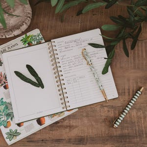 May include: A spiral-bound plant care journal with handwritten notes about eucalyptus, including scientific name, date, and care instructions. A floral patterned pen rests on the open page, while a green and white striped pen lies nearby.