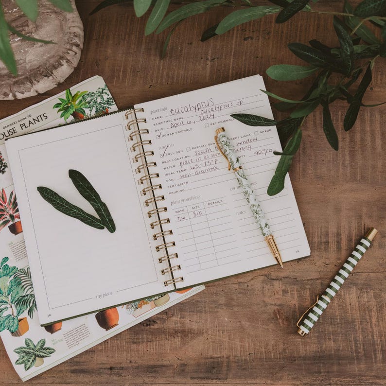 May include: A spiral-bound plant care journal with handwritten notes about eucalyptus, including scientific name, date, and care instructions. A floral patterned pen rests on the open page, while a green and white striped pen lies nearby.