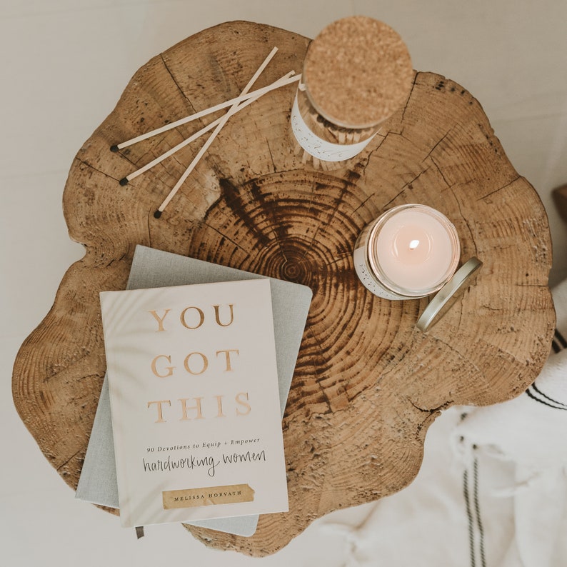 May include: A white book with gold lettering that reads "You Got This 90 Devotions to Equip & Empower Hardworking Women Melissa Horvath" on a wooden surface with a candle and matches.
