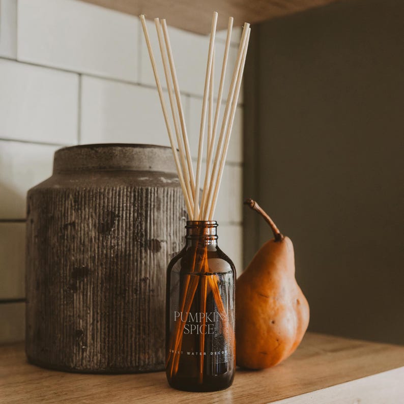 May include: A brown glass reed diffuser bottle with the text PUMPKIN SPICE printed on the front. The bottle contains several light-colored wooden reeds extending upwards. A textured gray cylindrical vase and a brown pear sit nearby.