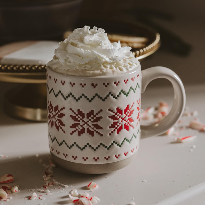 May include: A ceramic mug with a white base and a textured red and green Nordic-style snowflake pattern, filled with a hot beverage topped with a generous swirl of white whipped cream, surrounded by scattered small pieces of crushed peppermint candy.