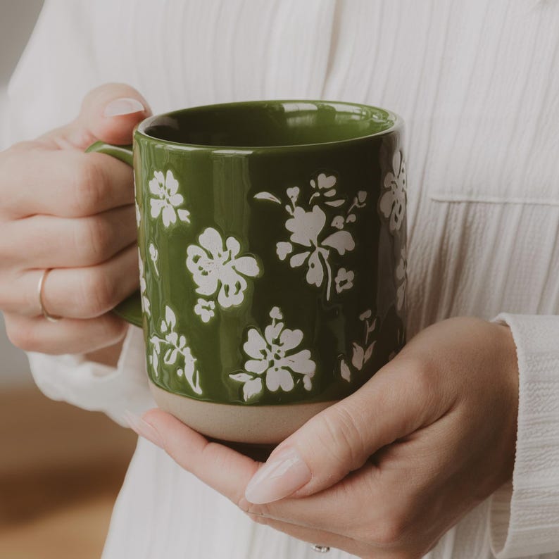 May include: A green ceramic mug with white floral designs. The mug has a beige base and a green handle. The mug is held by a person wearing a white shirt. The mug is a perfect addition to any kitchen.