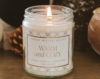 May include: A lit glass jar candle with a label that reads "WARM and COZY" with the scent notes of orange, cinnamon, clove, and pine. The candle is from Sweet Water Decor and made in the USA. The candle is in a clear glass jar.