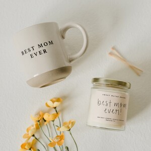 May include: A white ceramic mug with the text "BEST MOM EVER" and a beige bottom. A small glass jar with a gold lid contains a white candle with the text "best mom ever!" in black. The candle is labelled "Sweet Water Decor" and "Sea Salt, Jasmine, Wood & Cream". A bouquet of yellow flowers is in the bottom left corner of the image.