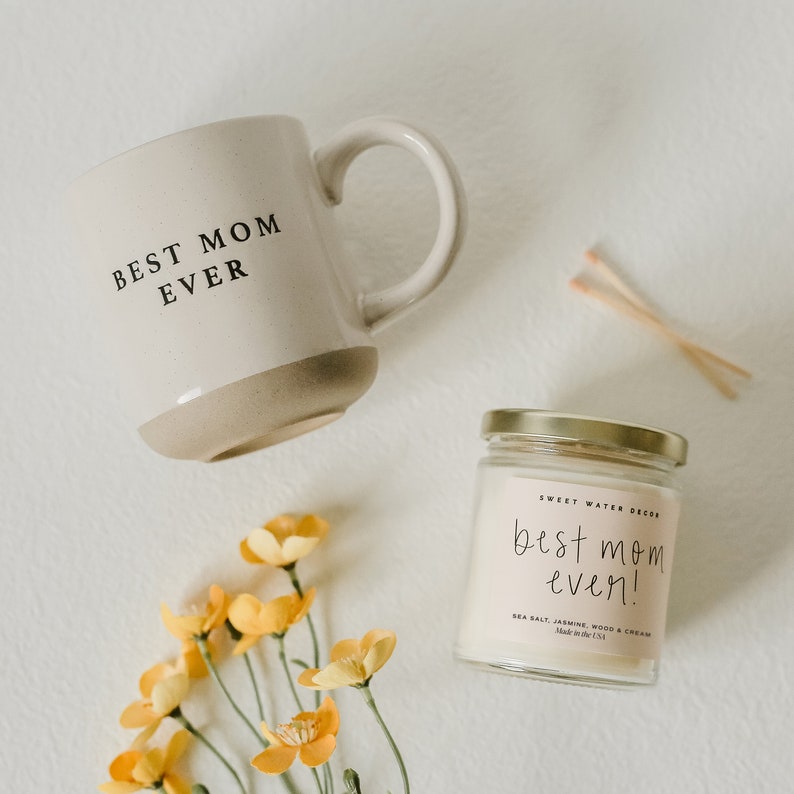 May include: A white ceramic mug with the text "BEST MOM EVER" and a beige bottom. A small glass jar with a gold lid contains a white candle with the text "best mom ever!" in black. The candle is labelled "Sweet Water Decor" and "Sea Salt, Jasmine, Wood & Cream". A bouquet of yellow flowers is in the bottom left corner of the image.