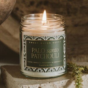 May include: A lit glass jar candle with a label that reads "PALO SANTO PATCHOULI" with the text "SWEET WATER DECOR". The candle is white with a gold and green label. The candle's scent includes clove, vanilla, patchouli, and cedarwood.
