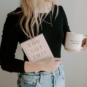 May include: A woman wearing a black long-sleeved shirt and blue jeans is holding a pink book with gold lettering that says "You Got This" and a white mug with the text "Be Still + Know".