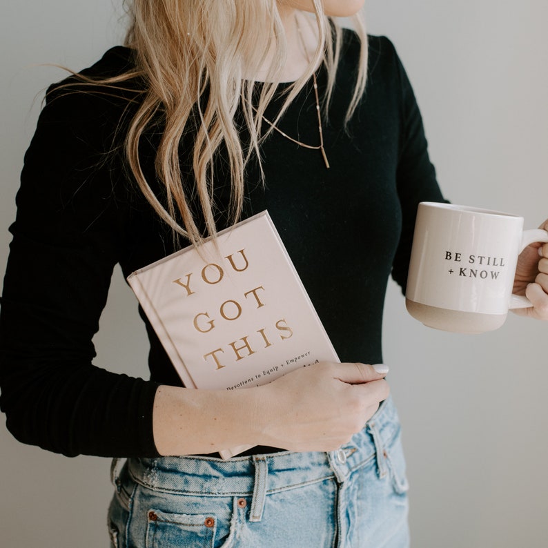 May include: A woman wearing a black long-sleeved shirt and blue jeans is holding a pink book with gold lettering that says "You Got This" and a white mug with the text "Be Still + Know".